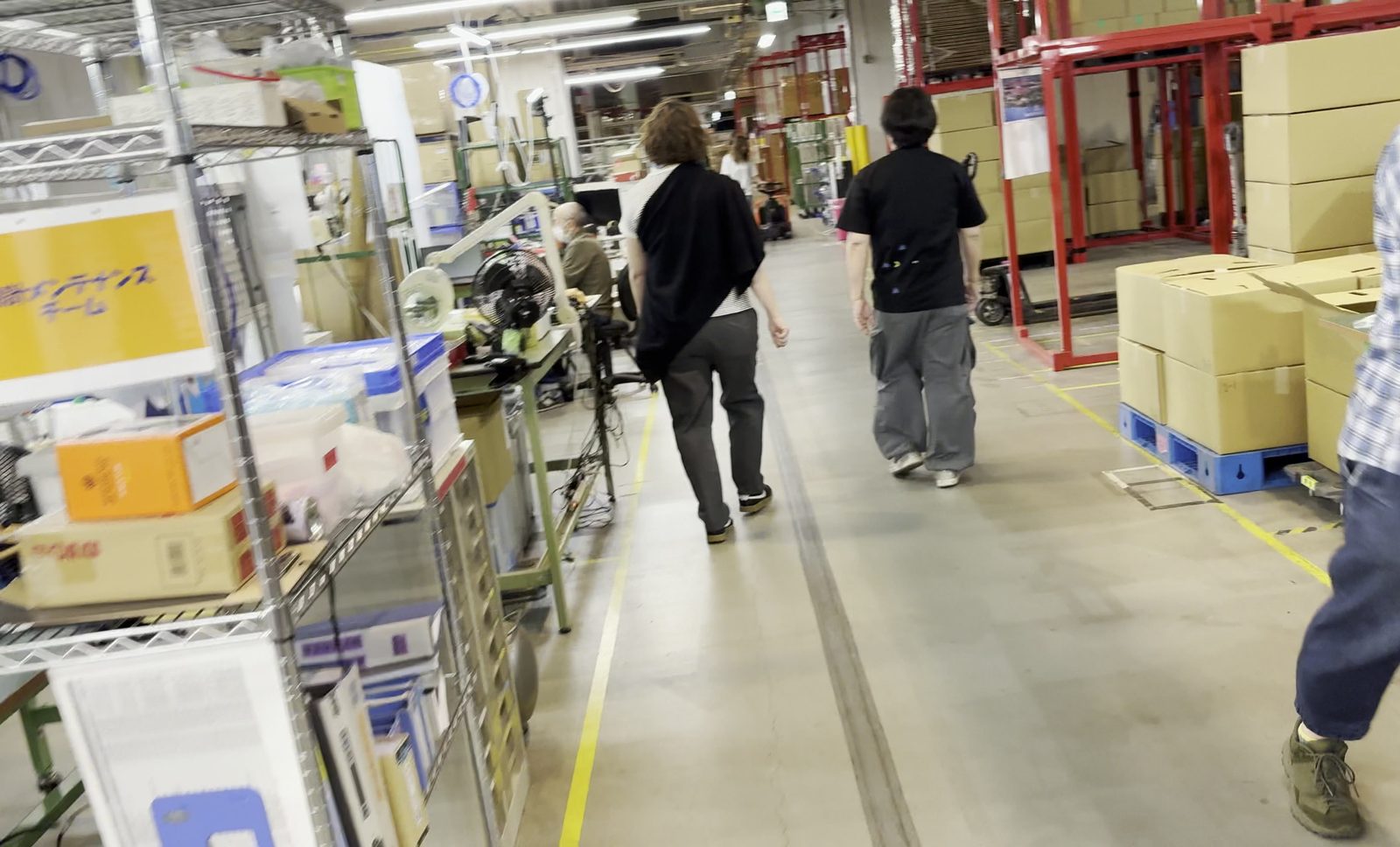 出品倉庫のフロア。作業台とラックが並び、奥にパレット積みの段ボールが見える